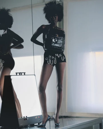 Model with voluminous dark hair wearing embellished black shorts and heels standing in a studio by Arnaud Lajeunie