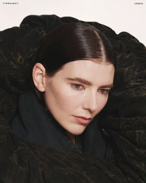 Close-up studio shot of a woman with dark hair in a dark green jacket and patterned black outerwear, looking down in a fashion style image.