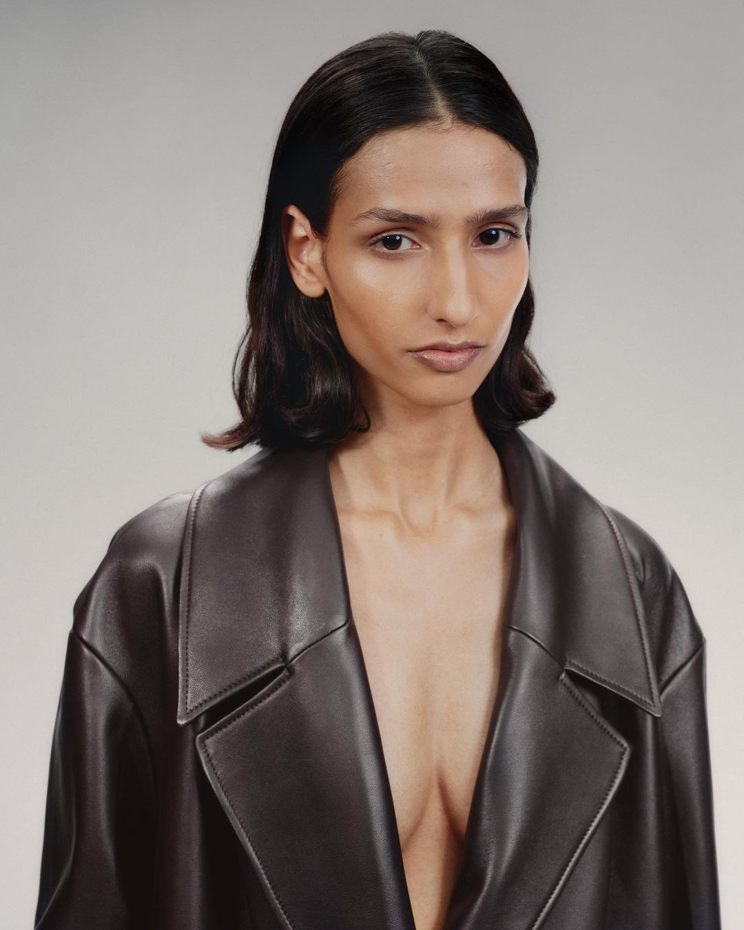 Portrait of a woman with medium-length dark hair wearing a dark leather trench coat against a neutral grey studio background.