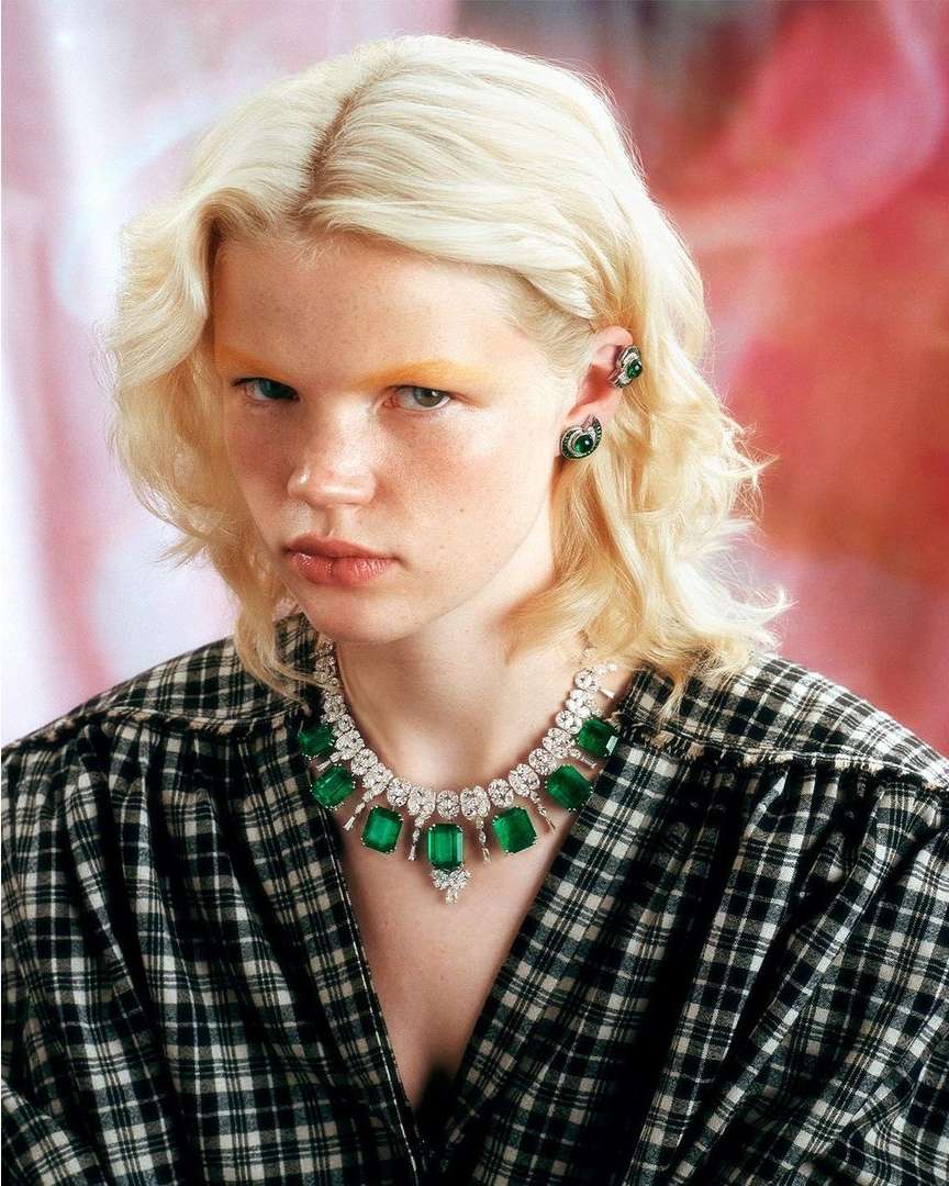 Close-up of a young woman with blonde hair wearing a black and white plaid shirt and a diamond and emerald necklace and earrings.