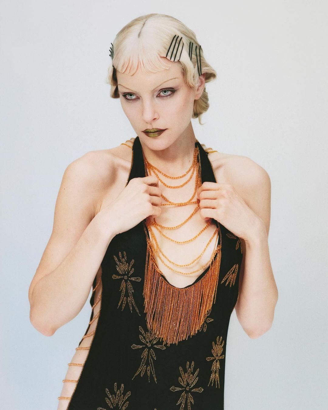 Woman in flapper style dress with blonde hair and gold fringed necklace posing in front of a light grey background in a studio.