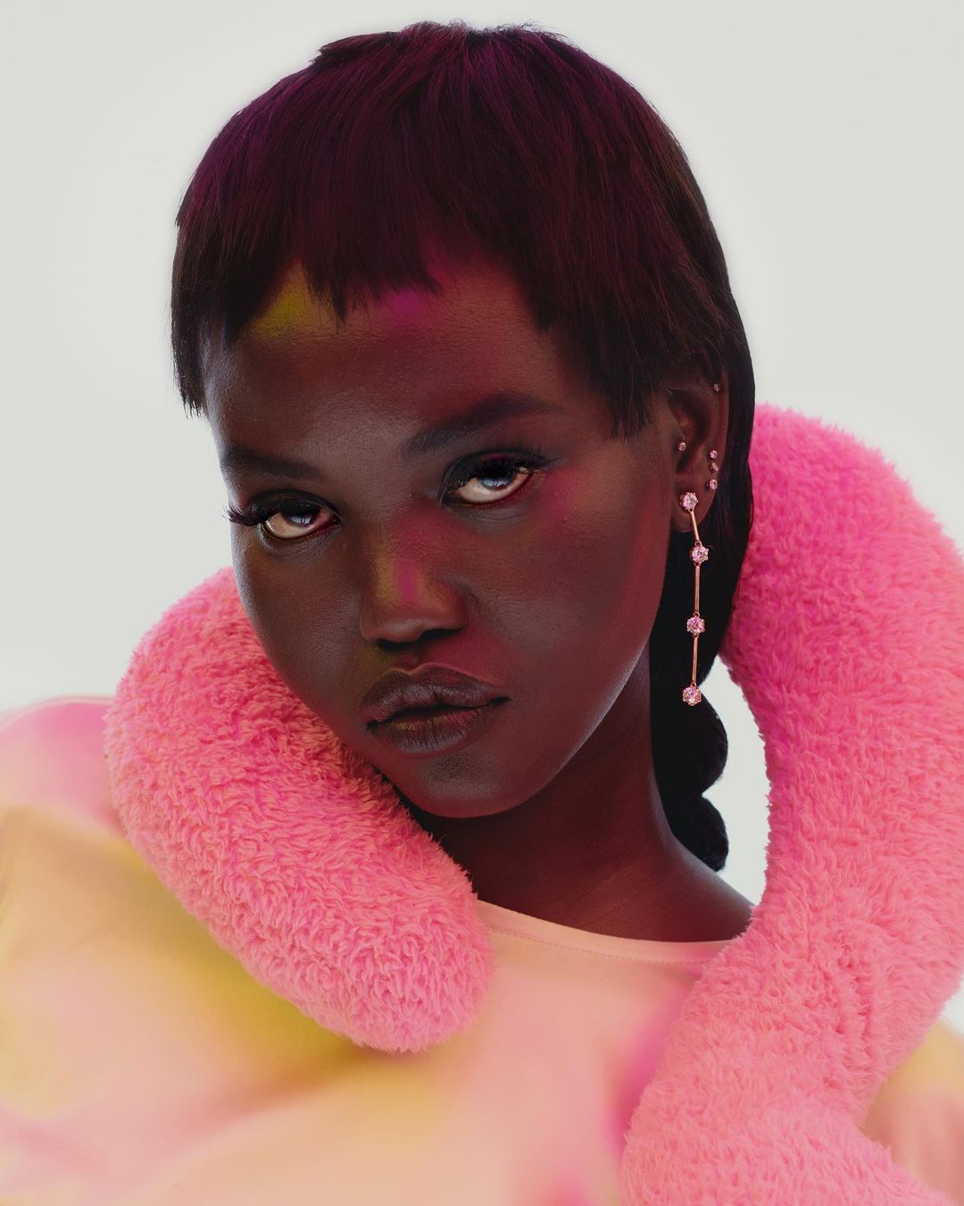 Close-up portrait of a young Black woman with short dark hair, wearing a pink fuzzy collar and drop earrings, against a white backdrop.