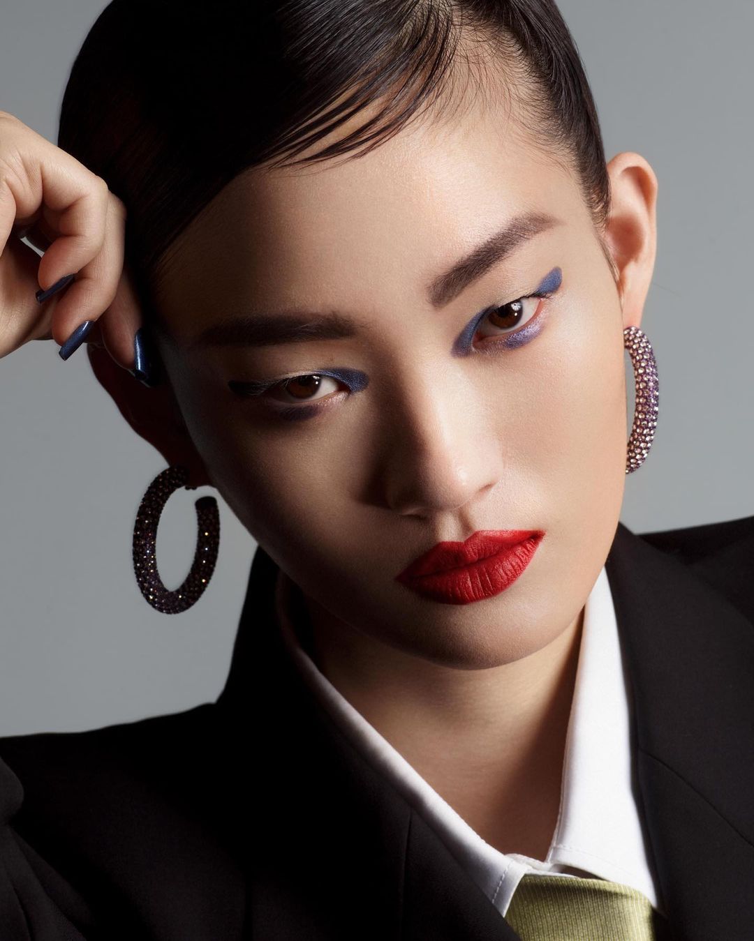 Close-up portrait of a woman with red lipstick, blue eye makeup, and jeweled hoop earrings by photographer Arnaud Lajeunie