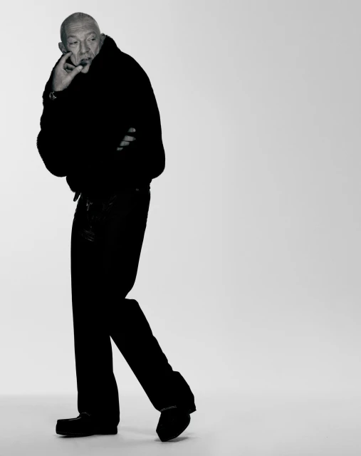 Studio portrait of a mature adult male in a dark outfit, standing in a relaxed pose with a contemplative expression, against a grey backdrop.