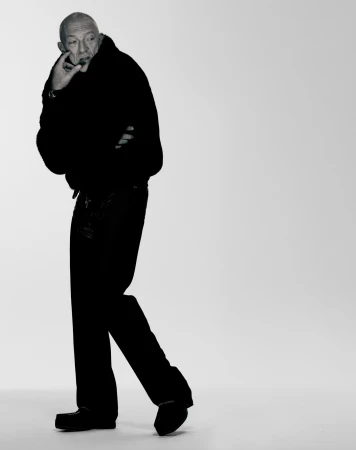 Studio portrait of a mature adult male in a dark outfit, standing in a relaxed pose with a contemplative expression, against a grey backdrop.