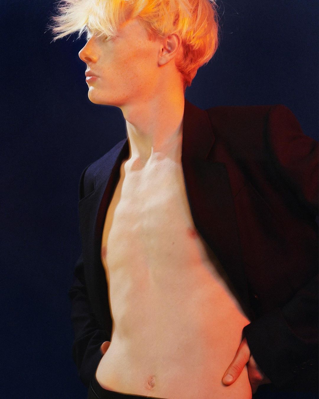 Profile of shirtless adult male with blonde hair wearing black blazer against dark blue backdrop, lit with a red/orange gel.