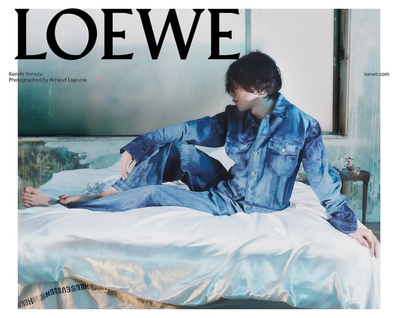 A person wearing a denim outfit lounges on a bed in a fashion editorial, alongside the Loewe brand logo and website address.