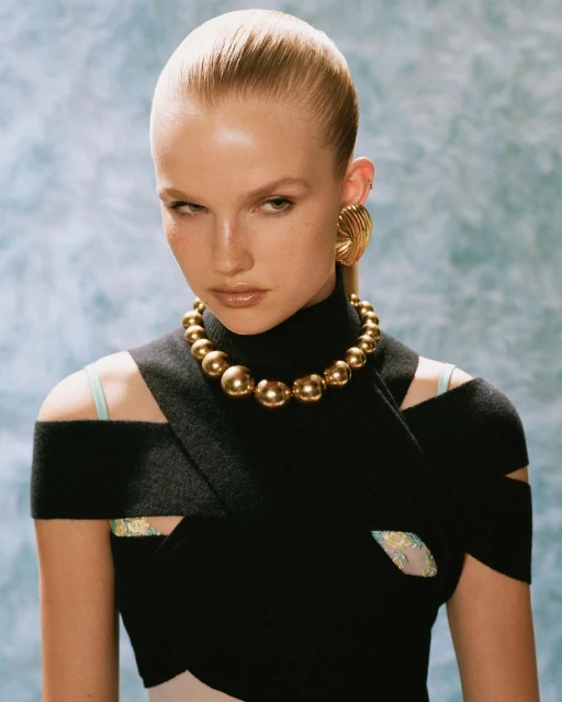 A fair-skinned woman in a high-fashion portrait wears a black dress and gold jewelry against a blue and white mottled backdrop.