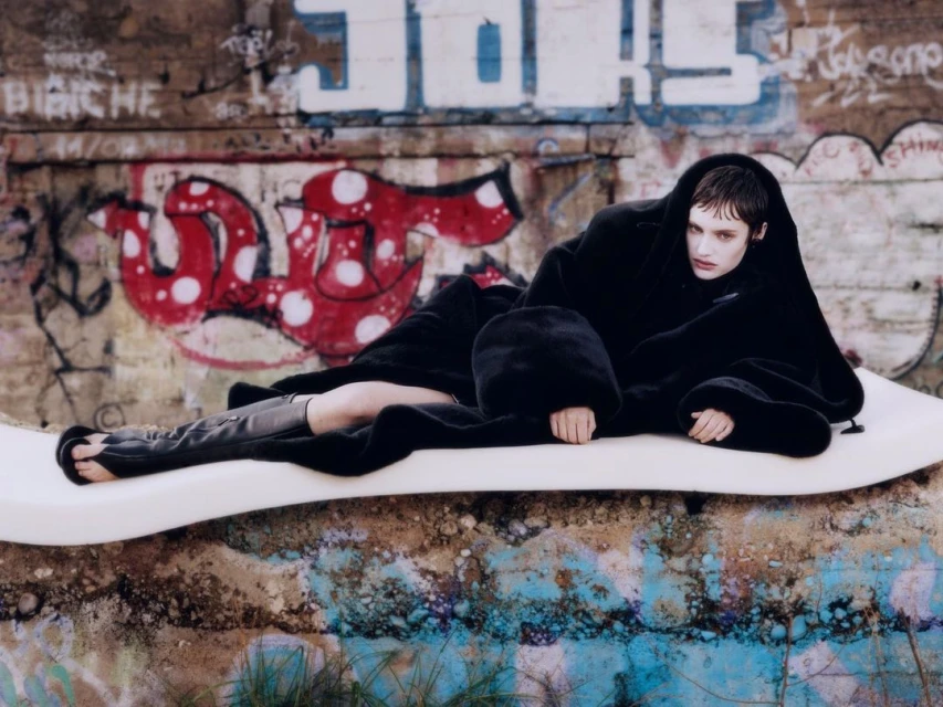 A woman in an oversized black coat poses outdoors against graffiti walls. She reclines on a white structure with her feet visible.
