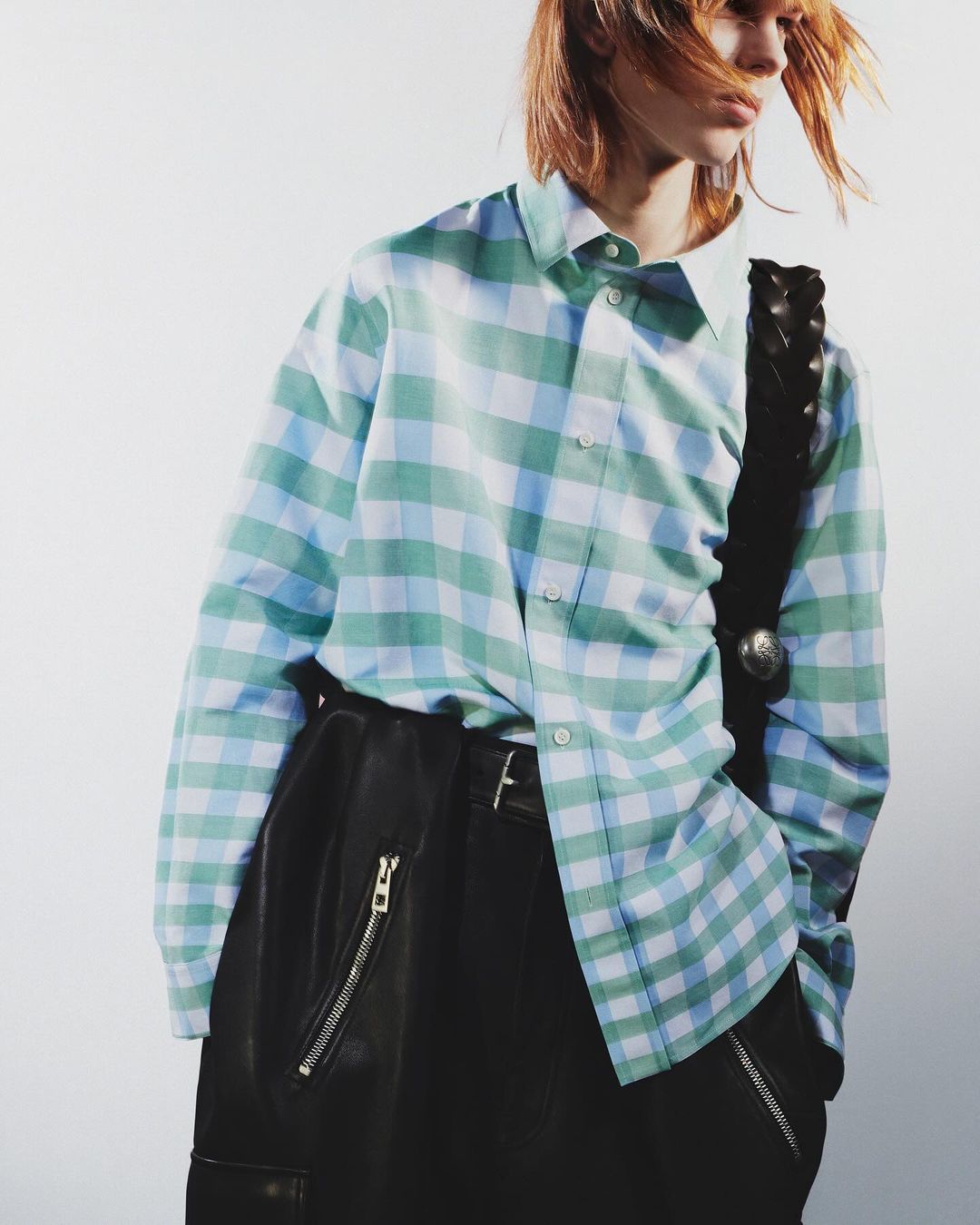 Fashion shot of person in an oversized plaid shirt, leather pants, and studded strap against a white background in a studio.