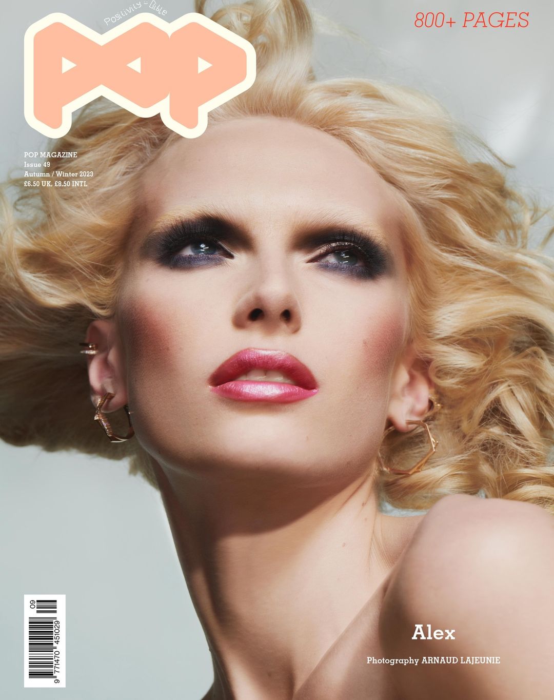 Close-up portrait of a blonde woman with dramatic smokey eye makeup and glossy pink lips against a plain background.