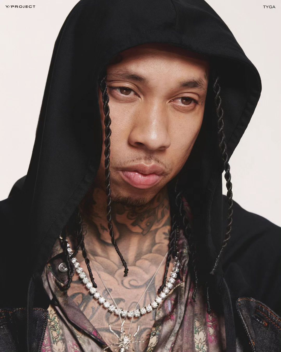 Close-up portrait of Tyga wearing a black hooded jacket and diamond necklaces against a bright backdrop. Braided hair is visible.