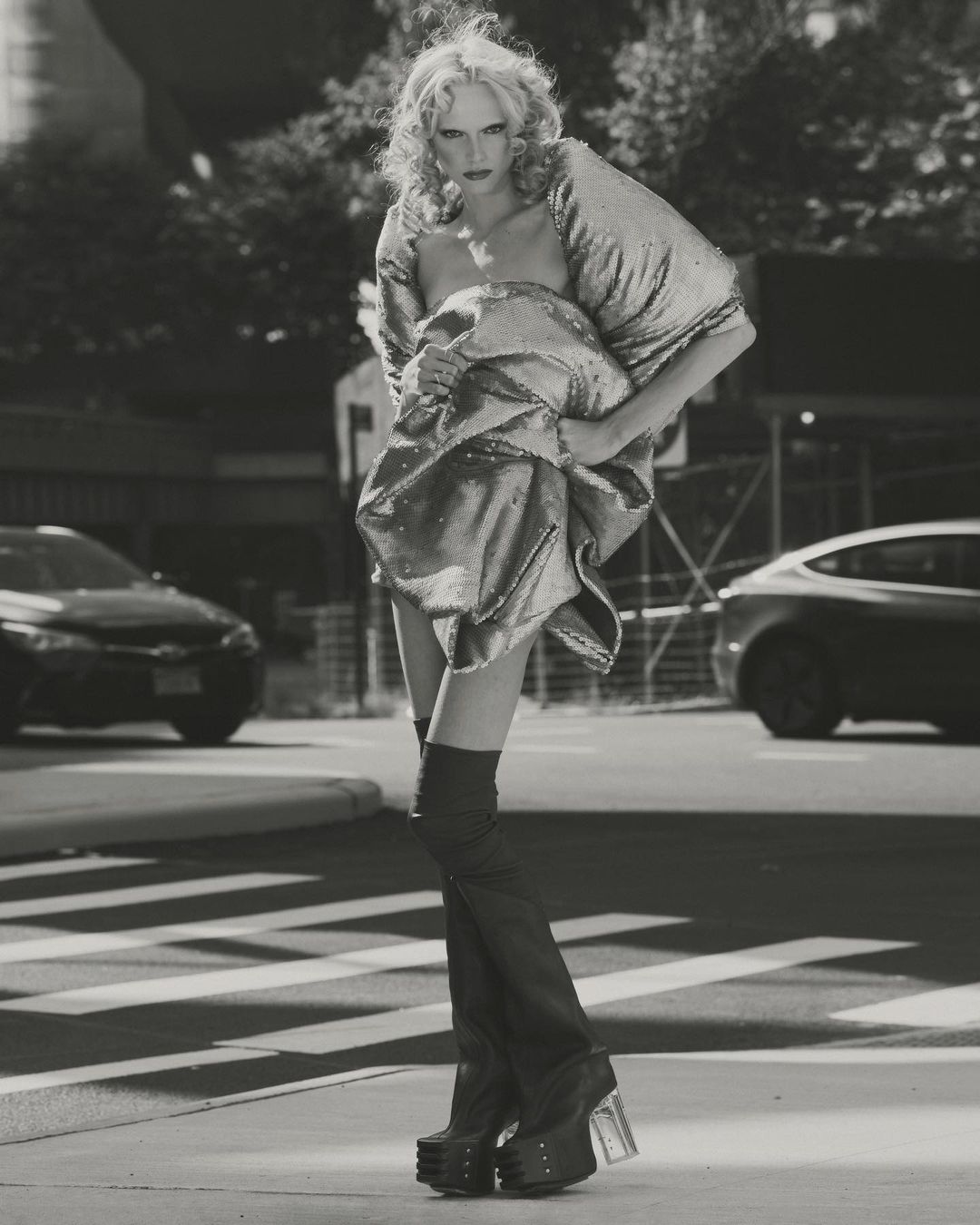 Monochrome fashion shot of a blonde model in a metallic dress and platform boots walking on a city crosswalk, with cars and buildings.