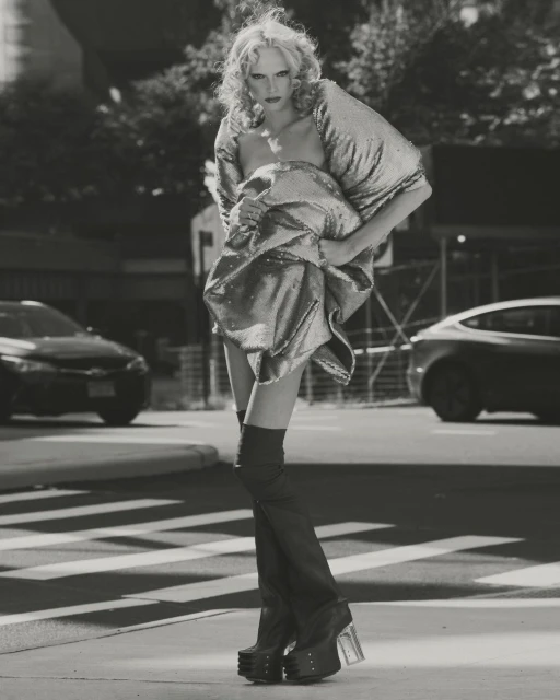 Monochrome fashion shot of a blonde model in a metallic dress and platform boots walking on a city crosswalk, with cars and buildings.