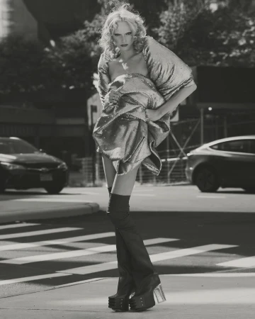 Monochrome fashion shot of a blonde model in a metallic dress and platform boots walking on a city crosswalk, with cars and buildings.