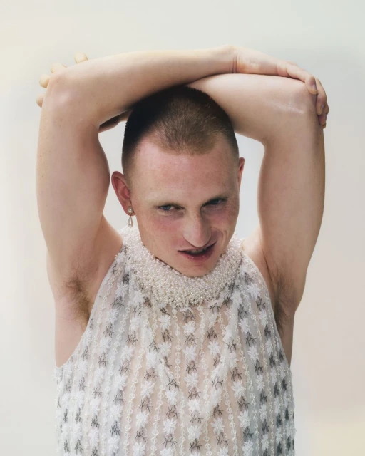 Person with shaved head in a sleeveless dress with pearl beaded collar and an earring, standing in front of a white background.