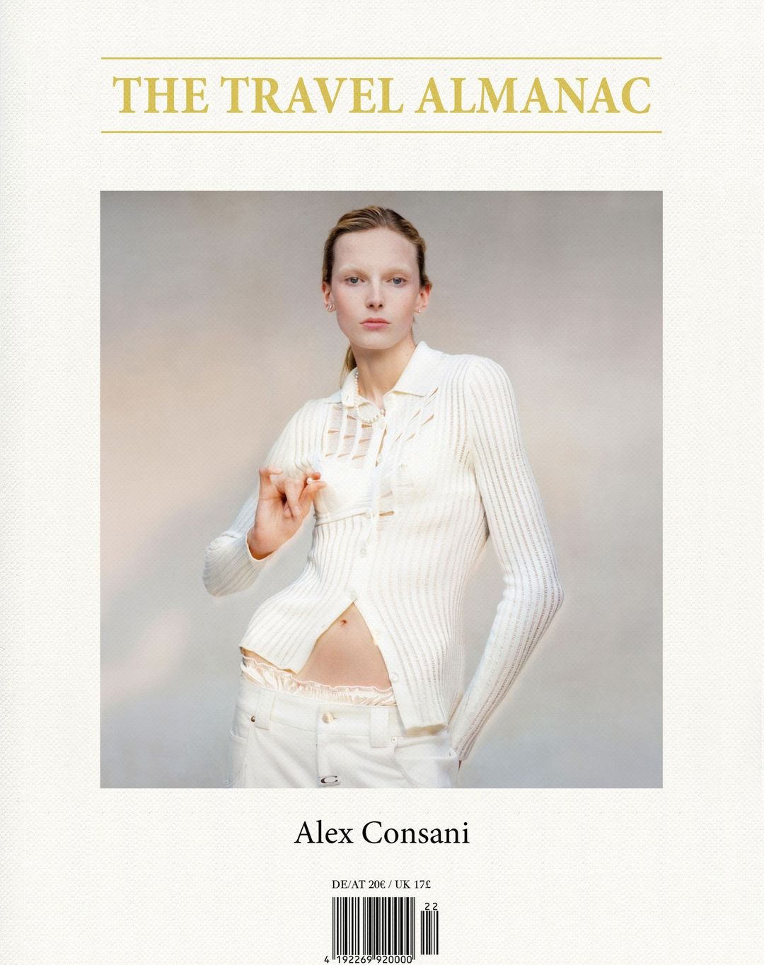 Fashion shot of a Caucasian female model in an off-white ribbed cardigan and white jeans holding a white card in a neutral indoor setting.