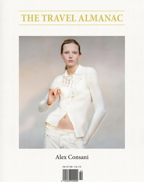 Fashion shot of a Caucasian female model in an off-white ribbed cardigan and white jeans holding a white card in a neutral indoor setting.
