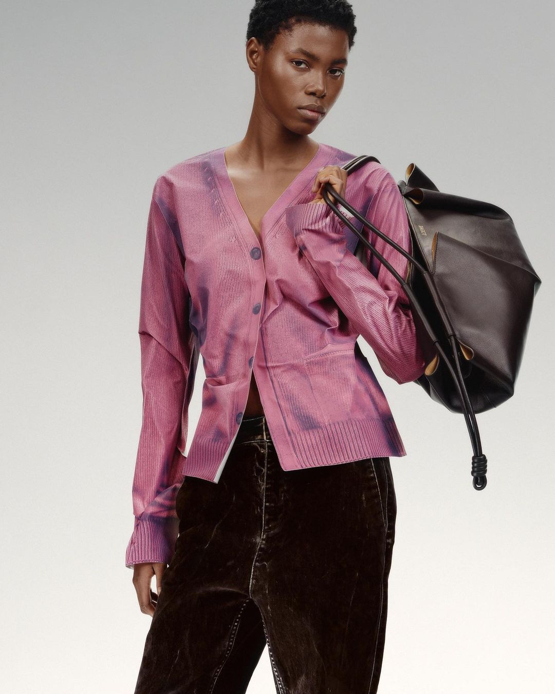 A young black woman in a pink cardigan and brown velvet pants, holding a black leather bag, poses against a soft, light-grey background.