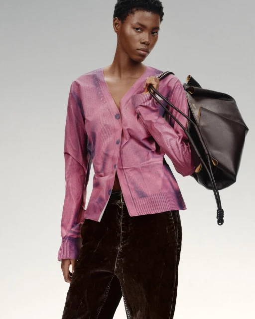 A young black woman in a pink cardigan and brown velvet pants, holding a black leather bag, poses against a soft, light-grey background.