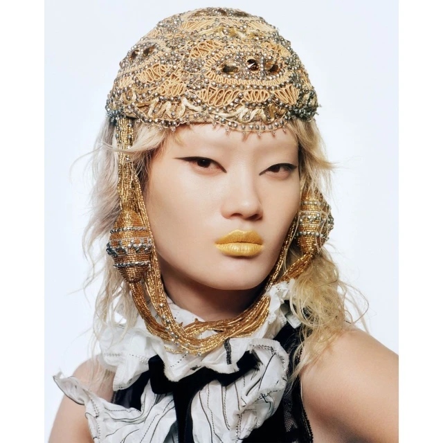 Eye-level studio shot of an Asian woman with pale skin, blonde crimped hair, heavy gold headpiece, and black and white dress against a white background.