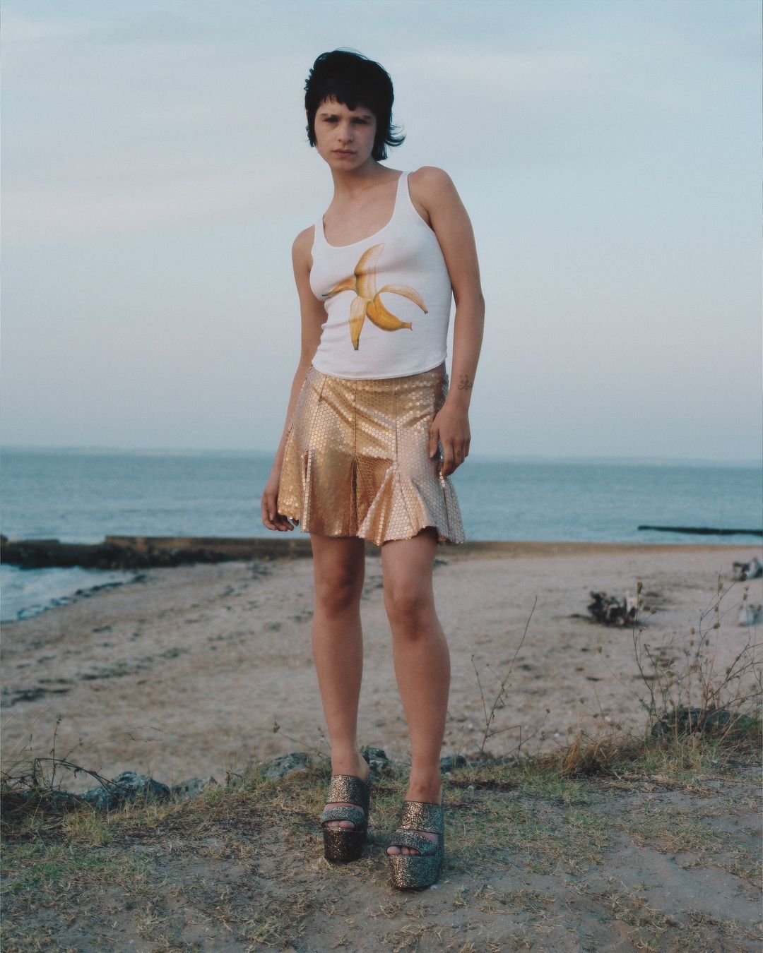 Woman stands on a sandy beach wearing a white tank top, gold skirt and platform shoes, with the ocean and sky in the background.