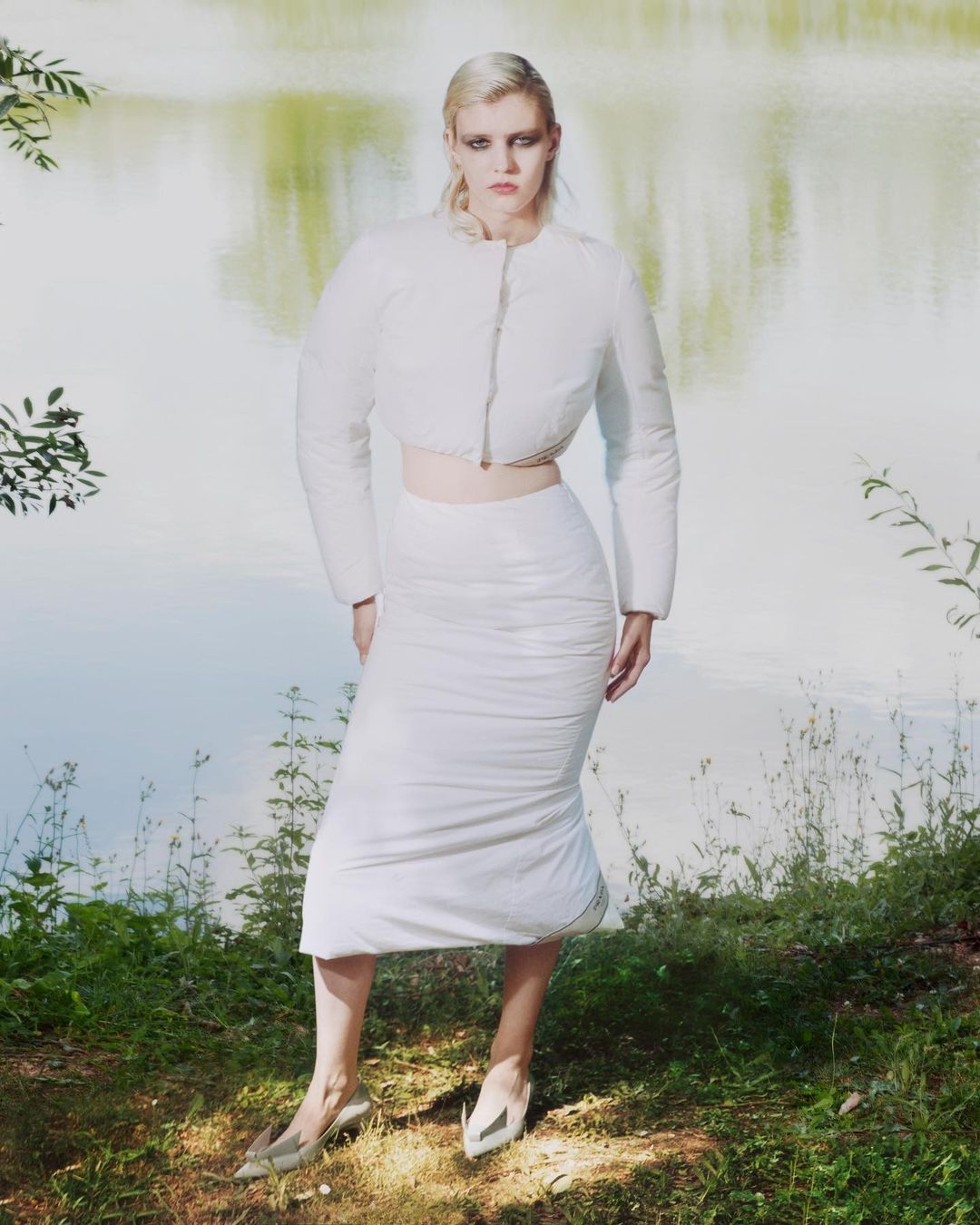 A woman in a white jacket and skirt stands outdoors near water, posing for a fashion shot with green grass and foliage surrounding her.
