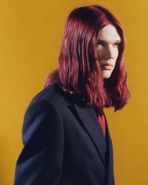 Portrait of a young adult male with long burgundy hair, wearing a black suit jacket against a solid yellow backdrop in studio lighting.