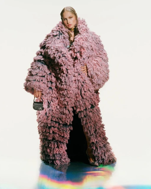 A stylish adult woman in a voluminous, textured pink coat stands indoors, holding a mini handbag, with a reflective, multicolored floor.