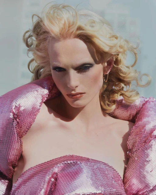 Close-up portrait of a model with blonde wavy hair wearing a sequined pink dress against a light blue background in an outdoor setting.