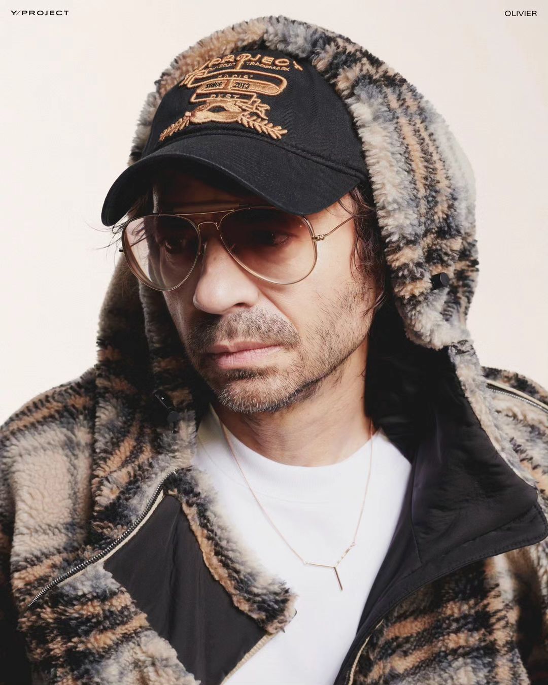 Adult man with aviator sunglasses and a cap, wearing a hooded jacket, poses indoors, with Y Project logo visible on the side.