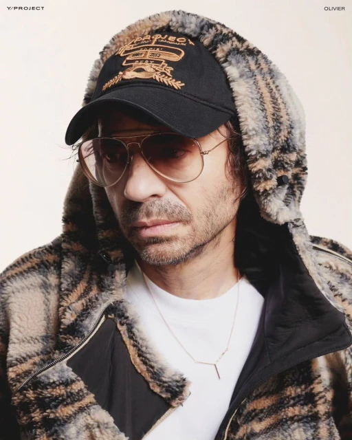 Adult man with aviator sunglasses and a cap, wearing a hooded jacket, poses indoors, with Y Project logo visible on the side.