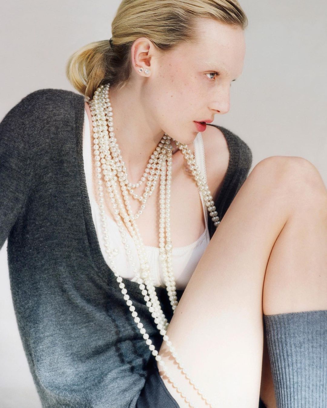Young adult woman with blonde hair wearing a grey cardigan, white tank top, and several strands of pearls, posing in a studio.