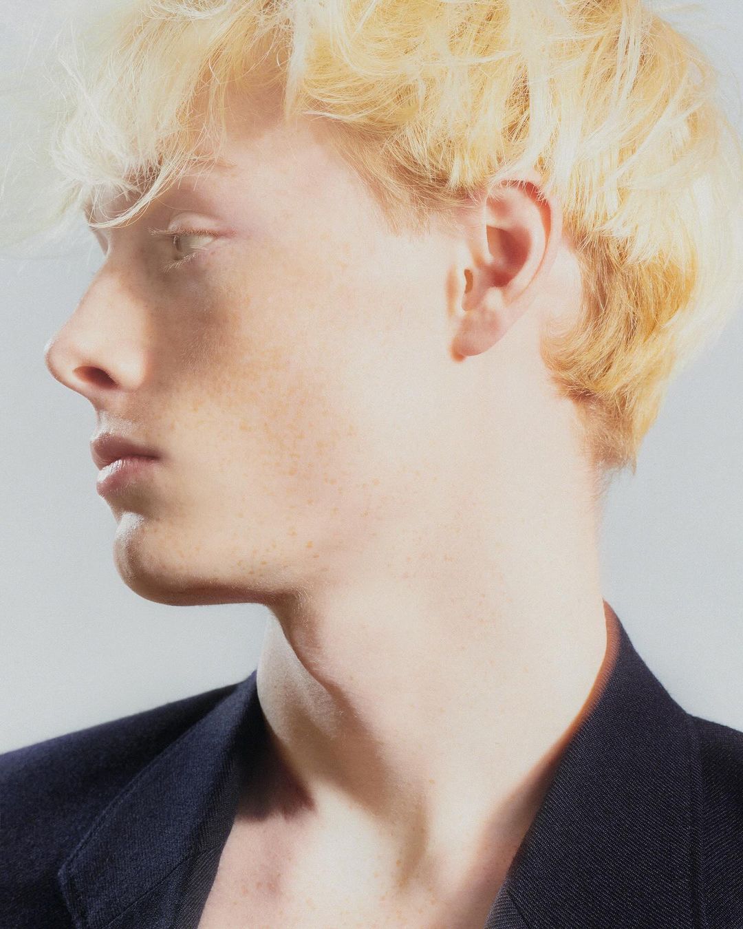 Profile portrait of a young man with blonde hair and a dark blazer against a plain light background by Arnaud Lajeunie