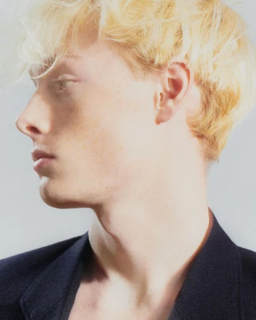 Profile portrait of a young man with blonde hair and a dark blazer against a plain light background by Arnaud Lajeunie