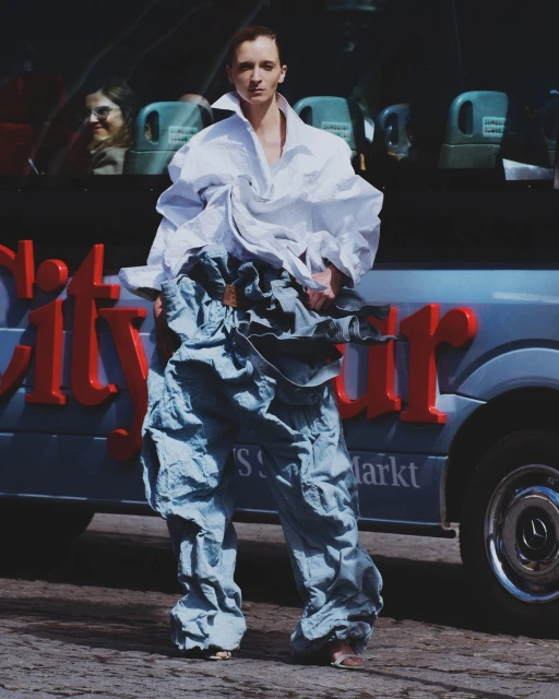 A fair-skinned adult female stands in a white shirt and layered light blue cargo pants in front of a City Tour bus.