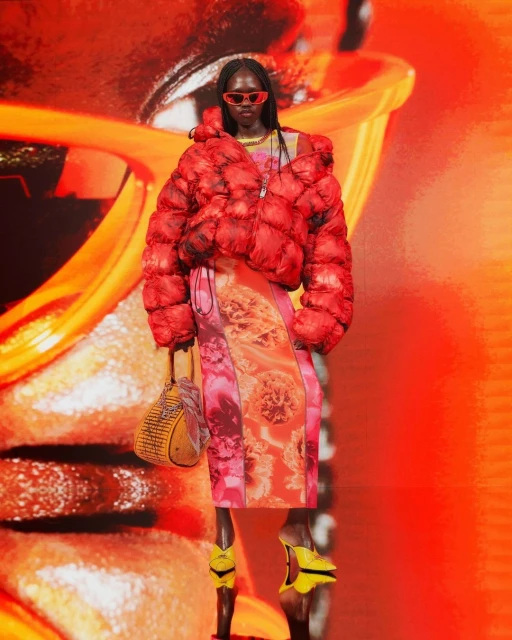 Fashion model on runway wearing puffer jacket, patterned dress, and sunglasses against a digitally enhanced orange background.