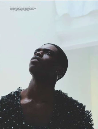 Portrait of a Black man looking upward, wearing a dark embellished top against a bright background.