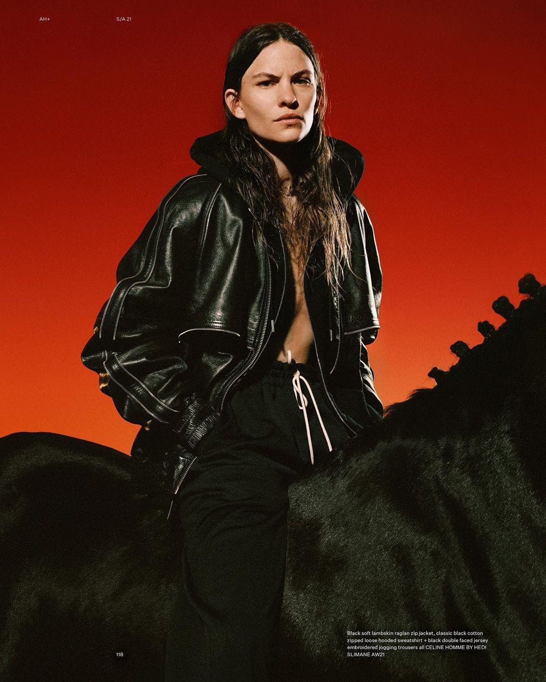 A woman with long brown hair poses confidently atop a black horse statue in an editorial shot against a gradient orange background.