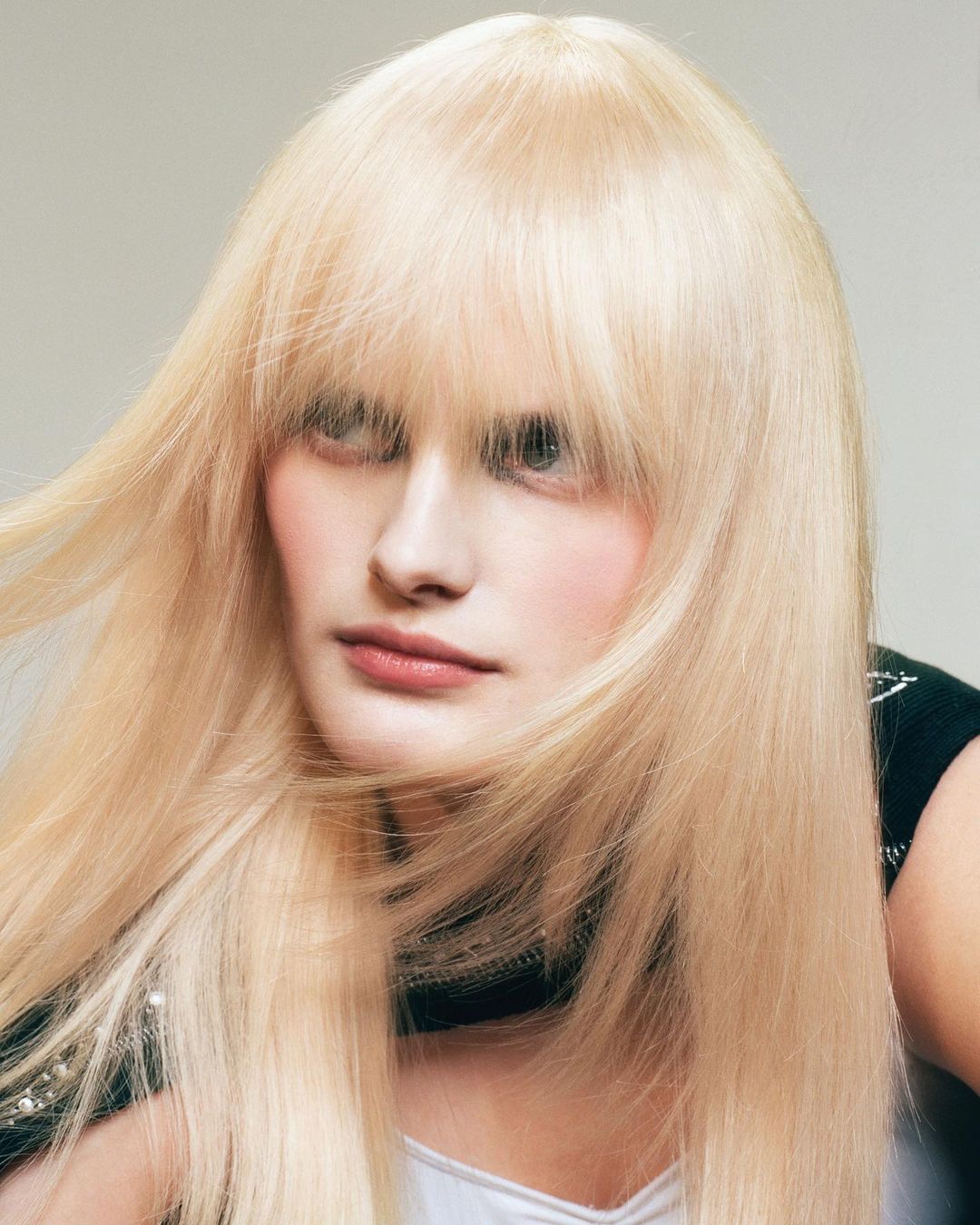 Portrait of a blonde woman with long hair and full bangs in a beauty and fashion shot, posing in front of a neutral background.