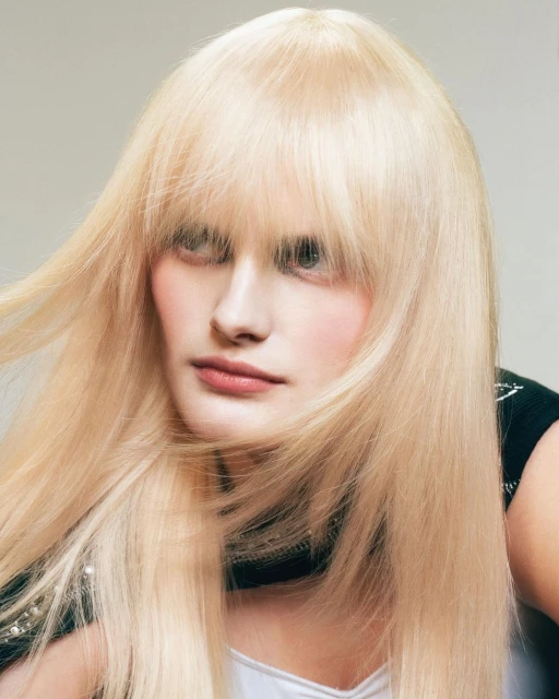 Portrait of a blonde woman with long hair and full bangs in a beauty and fashion shot, posing in front of a neutral background.