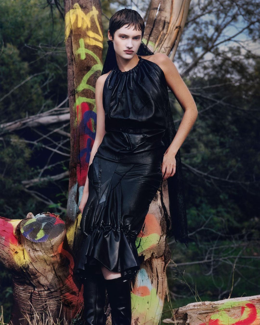 A model in black leather dress poses beside a tree with graffiti in an outdoor setting, creating a striking fashion editorial shot.
