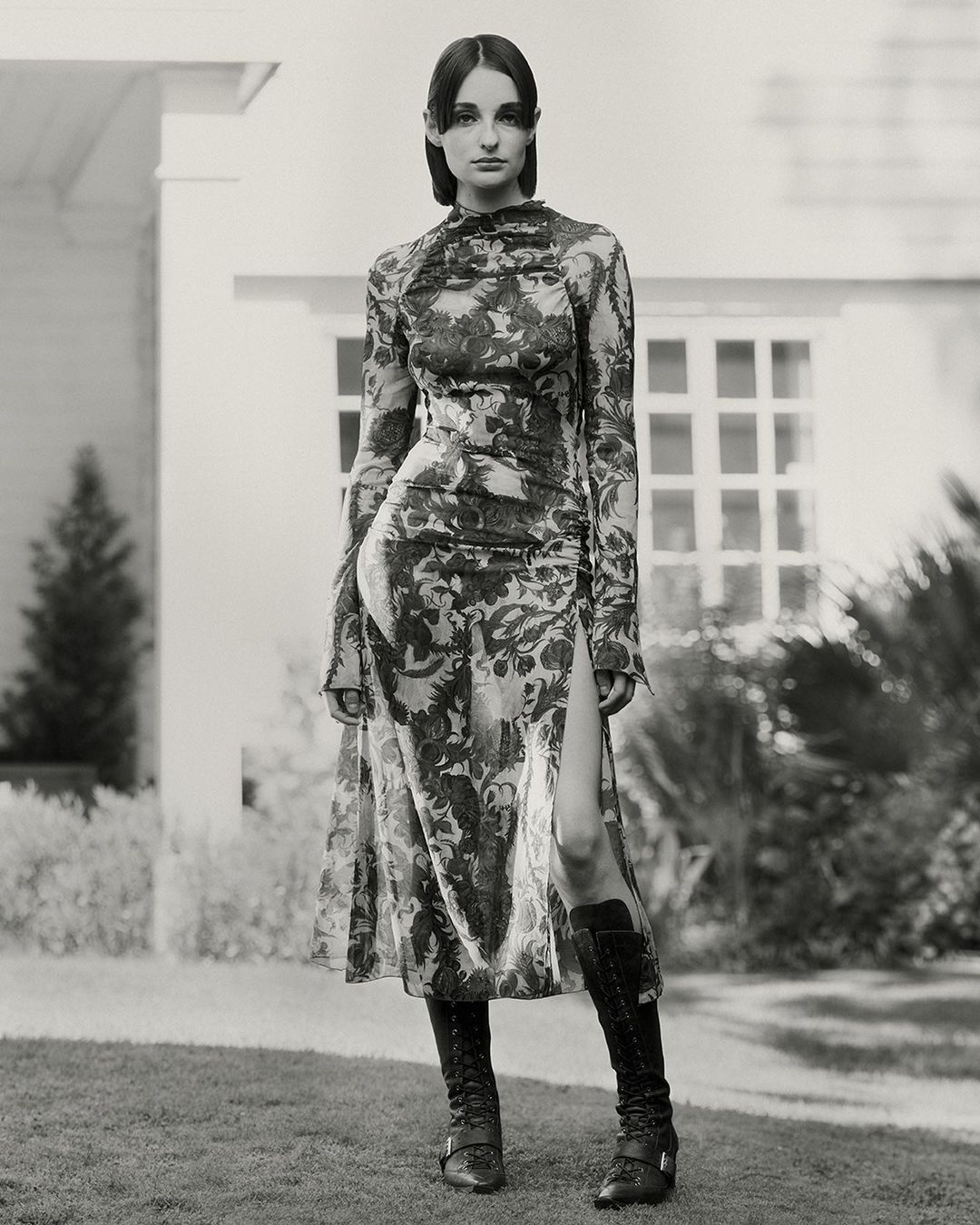A woman in a long-sleeved, floral midi dress and knee-high boots stands outdoors against the backdrop of a house and manicured lawn.