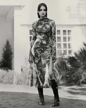 Model in a long floral print dress and tall boots standing outdoors in front of a white building with windows.