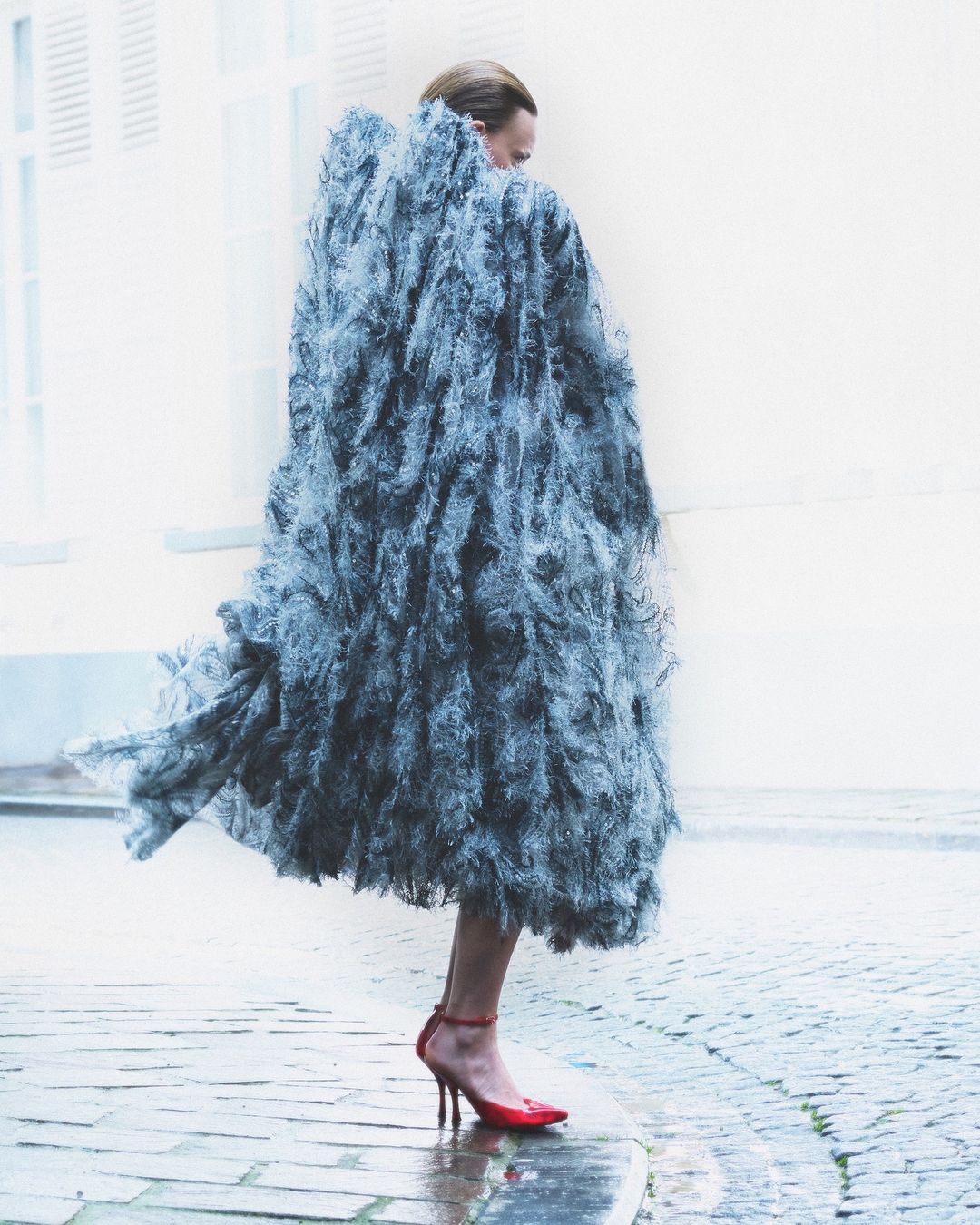 Model in a textured grey coat and red heels walks on a cobblestone street in a fashion editorial shot with natural outdoor lighting.