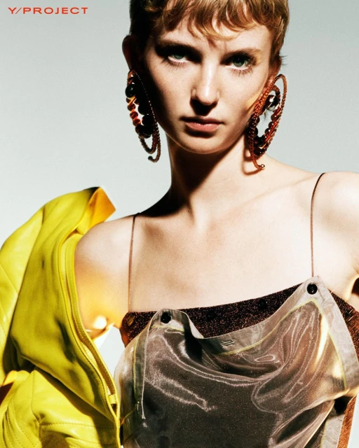 Close-up of woman with short blonde hair wearing large hoop earrings and a shiny silver top with a yellow jacket on her shoulder.