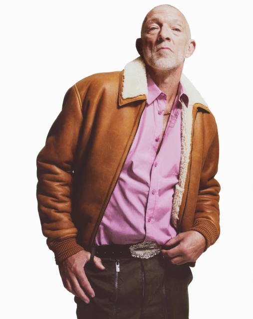 Portrait of adult man with beard, wearing pink shirt and brown jacket with fur collar, posing against a white backdrop.