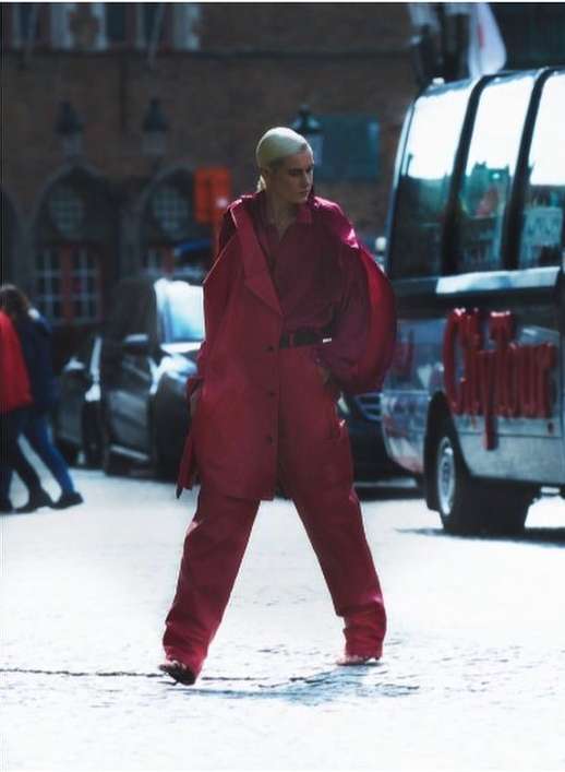 Fashion shot of an adult female model in a red coat and pants in an urban setting, walking on a cobblestone street near vehicles.