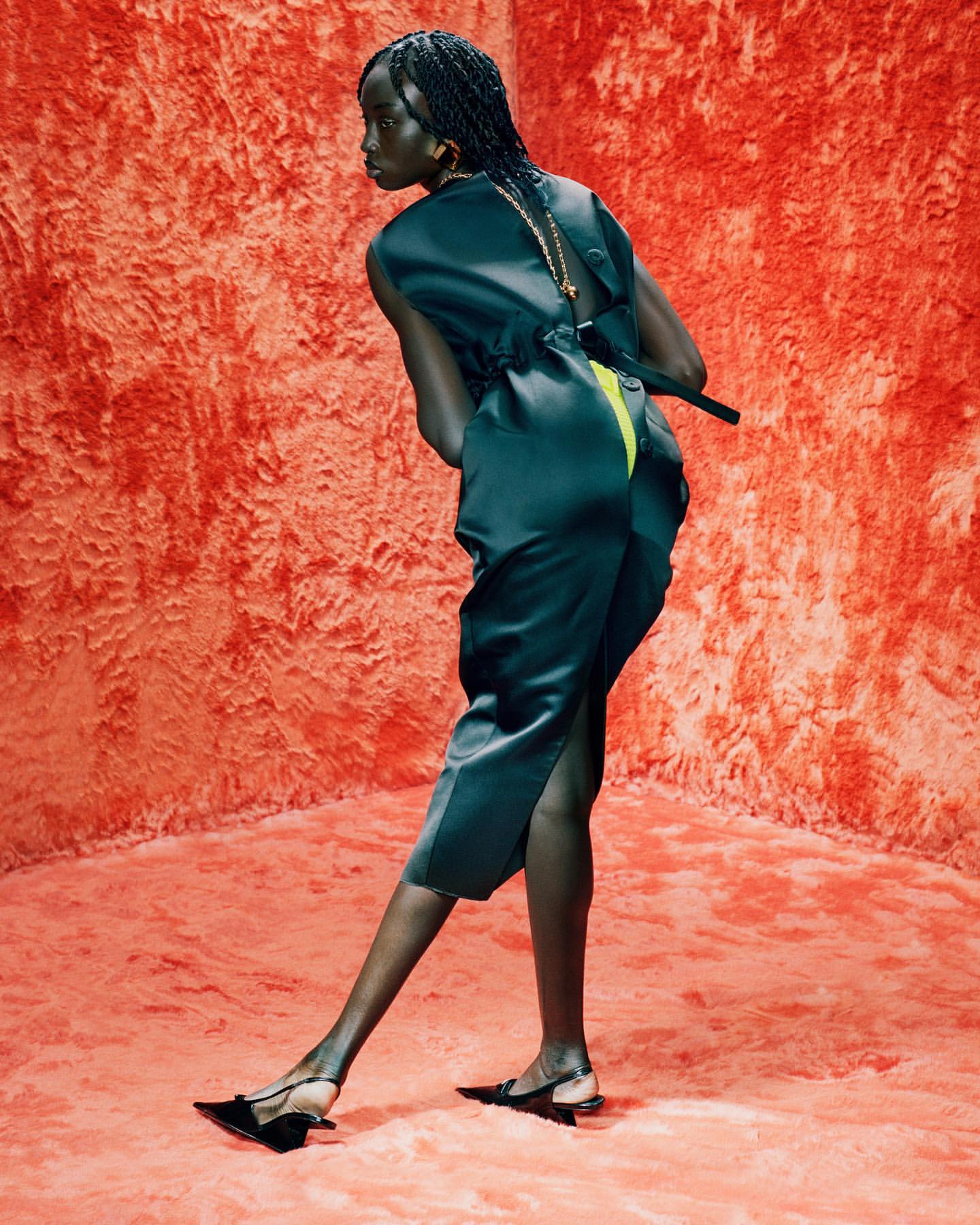 An African woman in a dark green sleeveless jumpsuit with braided hair poses in a textured orange studio with matching carpet.