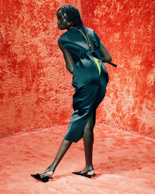 An African woman in a dark green sleeveless jumpsuit with braided hair poses in a textured orange studio with matching carpet.